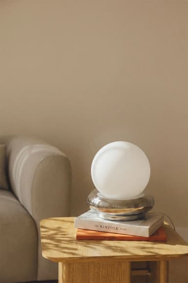 A modern table lamp featuring a spherical frosted white glass diffuser resting within a polished chrome base with a rounded, toroidal shape. The minimalist design emphasizes geometric forms and a sleek, reflective finish on the base, providing ambient light through the globe. — 3D model