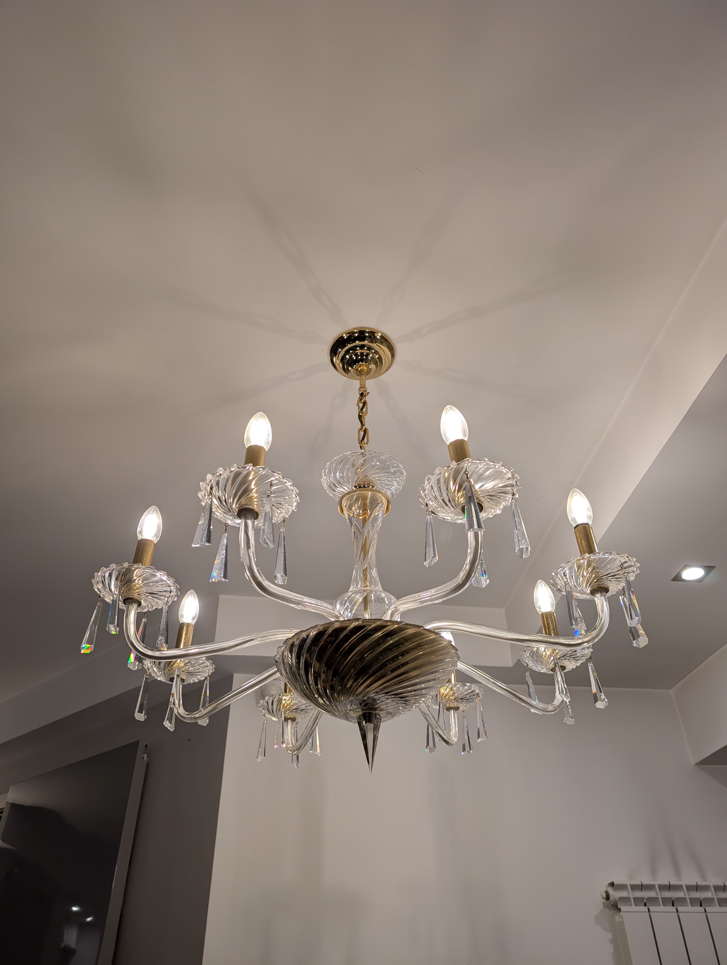 A multi-tiered chandelier with eight arms, featuring clear glass elements and crystal pendants. The central body has a twisted glass design, and the arms are curved, supporting candle-style light bulbs. The hardware appears to be gold-toned. — 3D model