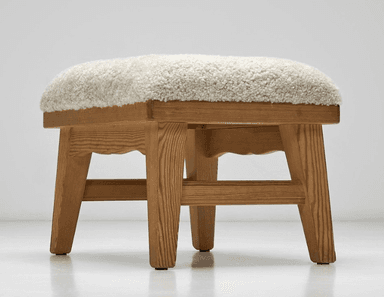 A rectangular stool featuring a thick, plush, cream-colored sheepskin upholstery top over a natural light-toned wooden frame. The frame has visible grain, sturdy splayed legs, and a subtly scalloped apron detail on the long sides. — 3D model