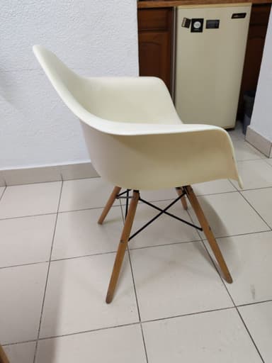 An off-white, molded plastic armchair with integrated armrests and a 'Eiffel Tower' style base consisting of splayed wooden dowel legs reinforced with black metal cross-supports. The chair is positioned on a light-colored tiled floor. — 3D model