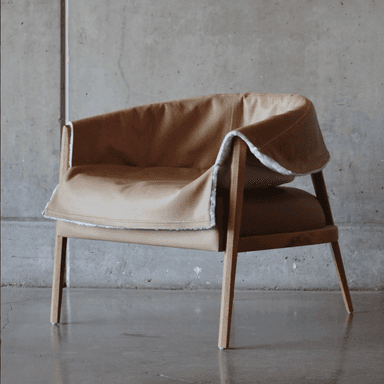 A contemporary armchair with a distinctive silhouette. The frame is constructed from light-colored wood, featuring four tapered legs and an open arm structure. The seat and back are upholstered in what appears to be tan leather, complemented by a plush, off-white sheepskin or faux shearling cover that drapes over the back and arms, creating a cozy aesthetic. The design suggests a blend of minimalist structure with textural richness. — 3D model