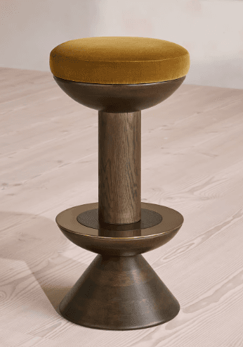 A contemporary bar stool featuring a round, thickly padded seat upholstered in gold/ochre velvet. The seat is supported by a dark, possibly walnut-toned, wooden column that widens towards the bottom into a conical base. A thin metallic ring, appearing to be brass or bronze, accents the transition point between the column and the base. — 3D model