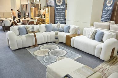 A large, modular sectional sofa featuring a curvilinear design, upholstered in a textured off-white bouclé fabric. It consists of multiple individual seating segments that create a curved arrangement. The set includes an integrated round wooden side table, a rectangular wooden end piece that serves as a surface or storage, and a separate matching upholstered ottoman. Several accent pillows in light blue and a geometric pattern are placed on the sofa. — 3D model