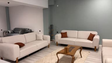A three-seater sofa with a light-colored, textured fabric upholstery. The sofa features a low profile with distinct seat cushions and a single-piece back cushion divided into three sections. It has a wooden frame and legs, with a visible wooden base extending around the perimeter. A single, small, rust-colored decorative pillow is placed on the left side of the seating area. — 3D model