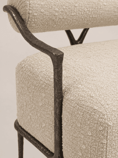An accent chair featuring a dark, hammered or forged metal frame and upholstered in a light-colored, textured boucle-like fabric. The design includes a separate cylindrical bolster for the backrest and a rectangular cushion for the seat. The metal frame forms minimal armrests and sturdy, tapered legs with small feet. The overall aesthetic is modern and artisanal. — 3D model