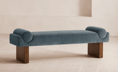 A modern daybed featuring a long, rectangular upholstered cushion with cylindrical bolsters integrated into each end. The seating surface is covered in a plush teal/slate blue velvet fabric. It rests upon two substantial, rectangular block legs made of dark-toned wood, characterized by a fluted or reeded texture on their outward-facing sides. The design is backless, emphasizing a sleek, contemporary profile. — 3D model