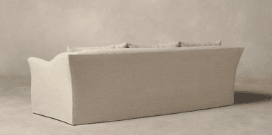 A three-seater sofa, viewed from the rear and side. The sofa features a loose slipcover construction with a skirted base that extends to the floor. It has a low profile with a gently curved armrest visible on the left. The back is composed of multiple cushions, suggesting a comfortable and plush design. The upholstery appears to be a textured linen or linen-blend fabric. — 3D model