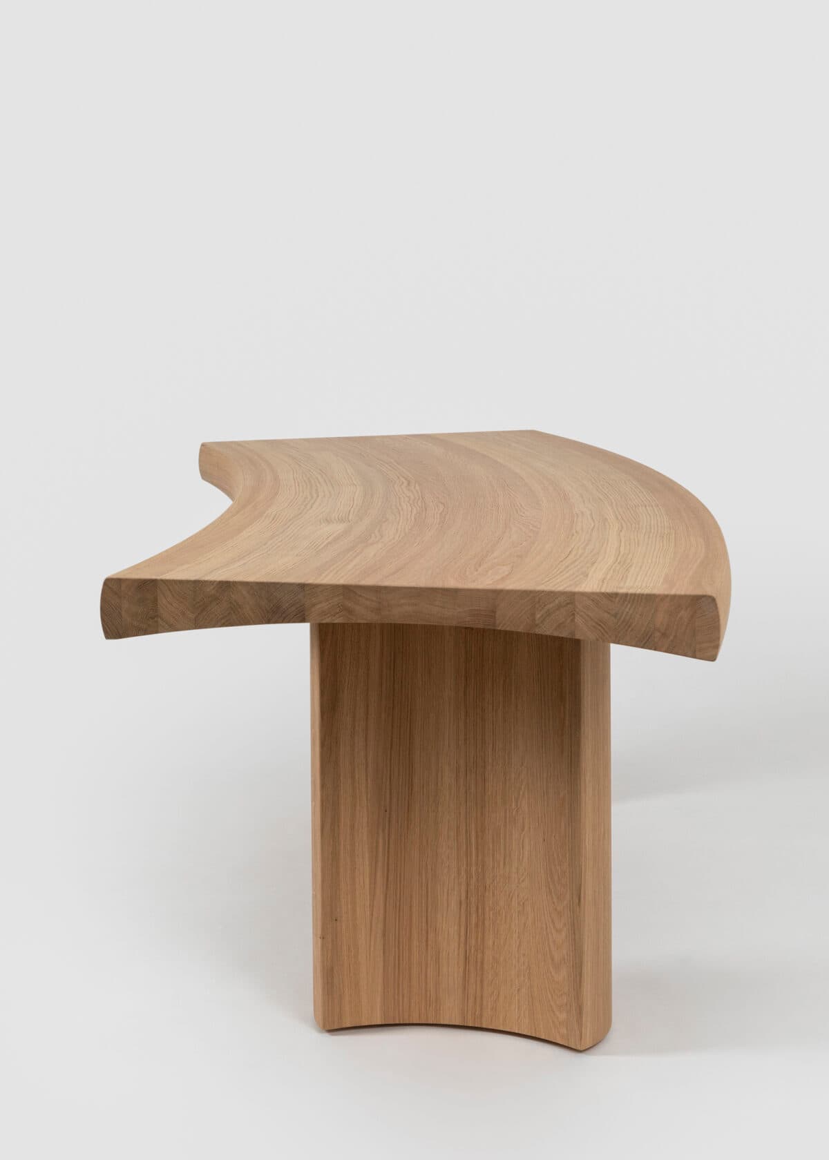 A modern curved bench crafted from light-toned solid oak wood. The design features a gently bowed top surface supported by two thick, substantial legs that are seamlessly integrated and follow the curvature of the top, creating a cohesive, sculptural form. The wood displays a natural grain, emphasizing its organic aesthetic. — 3D model