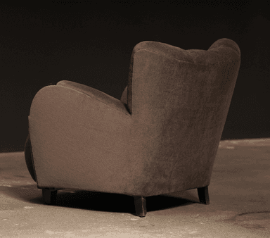 A robust, fully upholstered armchair viewed from the rear-right side, featuring a rounded backrest, integrated arms, and short, dark wooden legs. The chair is covered in a soft, dark taupe fabric. — 3D model