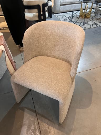 An upholstered dining chair featuring a contemporary sculptural form, integrated arms, and a continuous silhouette. It is covered in a light, textured, boucle-like fabric and is positioned at a dining table. — 3D model