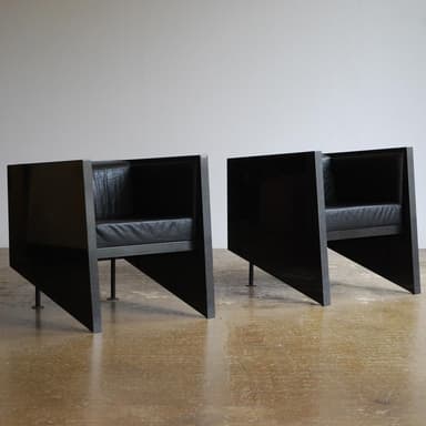 A pair of modern armchairs with a distinctive geometric design, featuring angular dark-stained wood or laminate frames and black leather seat and back cushions. The chairs present a minimalist and architectural aesthetic. — 3D model