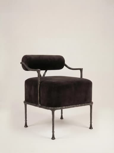 A robust armchair featuring a dark, possibly black or very dark brown, velvet upholstered seat and a cylindrical backrest. The frame is constructed from dark, textured, possibly forged iron or a similar heavy metal, exhibiting a somewhat raw and organic aesthetic. The legs are slender but robust, tapering slightly to small, disc-like feet. The backrest is suspended above the seat, supported by the frame's uprights, with a small V-shaped support element visible at the rear. — 3D model