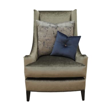 A fully upholstered wingback armchair, featuring a high back with subtly flared wings and rolled arms. It is covered in a textured, greenish-beige fabric, likely velvet or chenille. Decorative nailhead trim runs along the armrests and the bottom edge of the chair's frame. The chair rests on short, dark, tapered wooden legs. It is adorned with two accent pillows: one large rectangular pillow with a grey and beige abstract pattern, and a smaller solid navy blue square pillow with a central button tuft. — 3D model