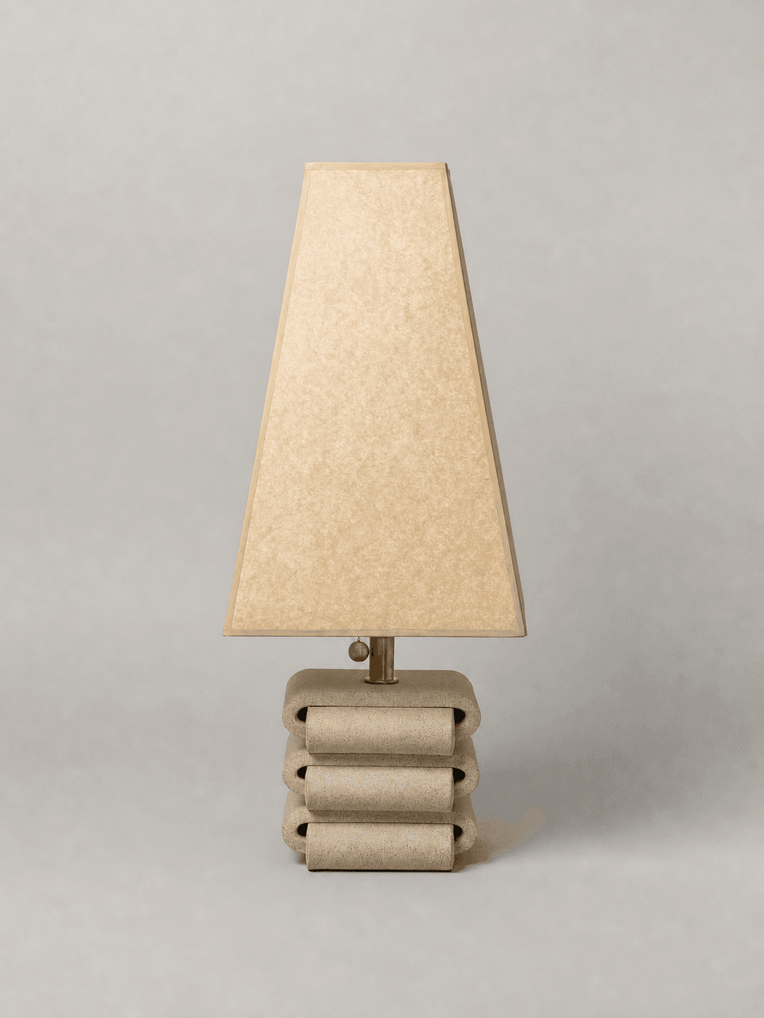 A table lamp with a trapezoidal lampshade and a stacked, rounded base. The base has a textured finish and appears to be constructed from multiple curved elements stacked vertically. The lampshade is a light, parchment-like color with a subtle texture. The fixture connecting the shade to the base is made of a metallic material, possibly brass. — 3D model