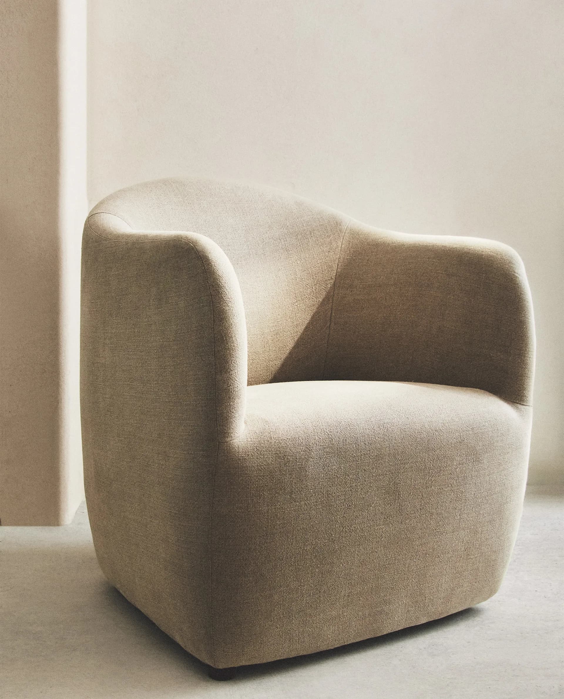 A contemporary upholstered armchair featuring a rounded, compact silhouette. The chair is entirely covered in a light beige, textured fabric, possibly boucle or a similar weave. It has a continuous backrest and integrated armrests that curve inward, creating a cocoon-like seating area. The base is subtly recessed, with small, dark glides barely visible beneath the main body. — 3D model