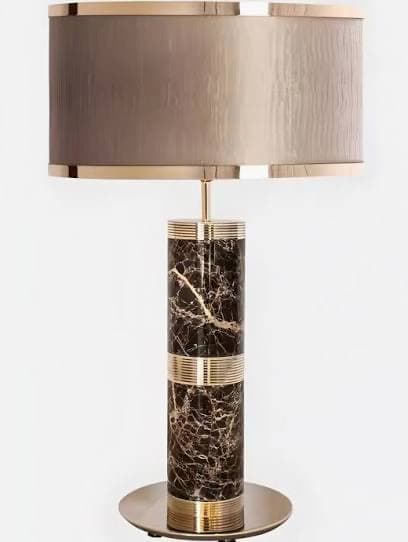 A contemporary table lamp featuring a tall, cylindrical base made of dark brown marble with prominent white and gold veining. The marble column is accented with ribbed brass bands. It is topped with a neutral-toned fabric drum shade, trimmed with polished brass on the upper and lower edges. A flat, circular brass plate forms the lamp's base. — 3D model