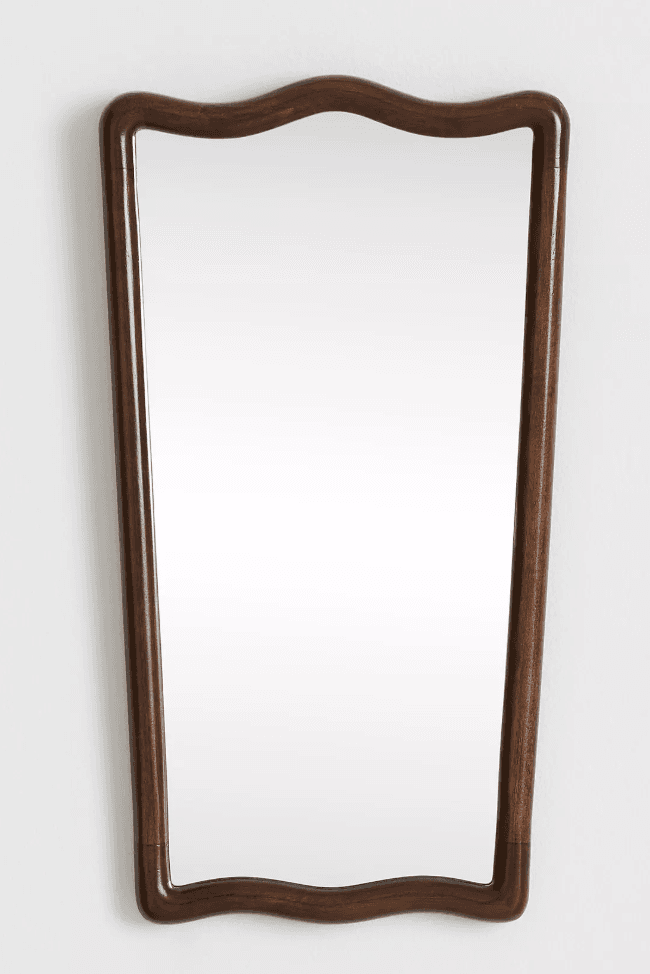 A wall-mounted mirror with a distinctively shaped frame. The frame exhibits a scalloped or wavy pattern along its top and bottom edges, while the sides are gently curved inwards, creating a somewhat hourglass silhouette. The material appears to be wood with a dark, polished finish. The mirror itself is rectangular with rounded corners and reflects a plain white background. — 3D model