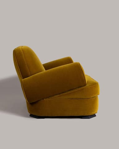 A compact, rounded armchair upholstered in a mustard-yellow velvet fabric. The armchair features a high, curved backrest that seamlessly transitions into padded armrests. The seat is low and broad, with a plain upholstered base. The overall design is plush and inviting, with a visible seam line indicating the upholstery construction. — 3D model