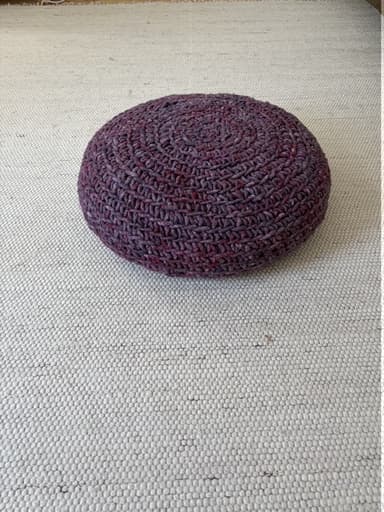 A round, disc-shaped floor pouf with a chunky, hand-crocheted or knitted exterior in a variegated dark reddish-purple or plum color. The textile features a prominent circular stitch pattern radiating from the center. — 3D model
