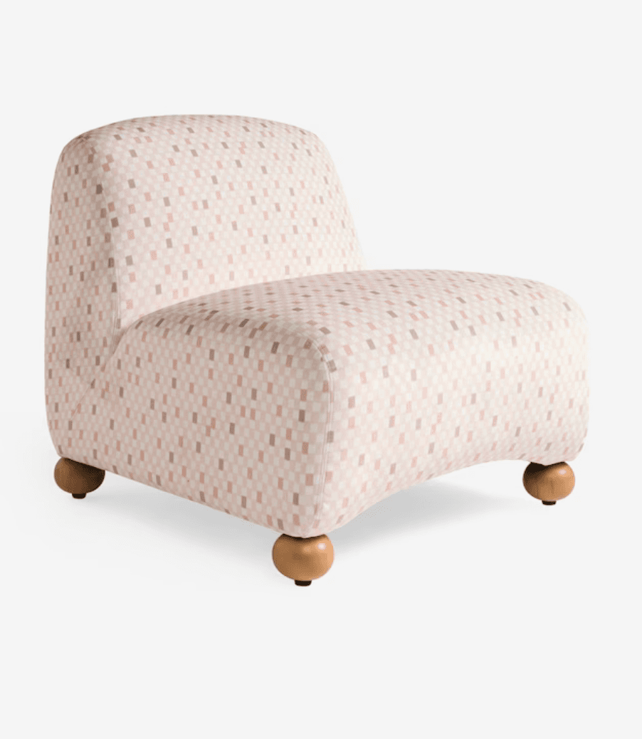 An armless upholstered chair featuring a contemporary design. The chair is covered in a fabric with a pixelated checkered pattern in shades of pink, beige, and light brown. It stands on four spherical light-toned wooden feet, contributing to its soft and rounded aesthetic. — 3D model