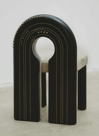 A contemporary chair with a distinctive U-shaped backrest that forms a cutout. The seat and the curved portion of the backrest are upholstered in a light beige, suede-like fabric. The frame is composed of dark, possibly black or dark wood, elements. The outer curved frame of the backrest and the vertical supports of the legs are adorned with a series of small, brass or gold-colored studs. The legs are thick and rectangular, with a dark finish. The chair's design is modern and somewhat sculptural, featuring a negative space in the upper backrest. — 3D model