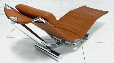 A contemporary chaise lounge with a curved, reclining design. The seat and back are upholstered in a stitched, padded orange-brown leather or leather-like material. The frame appears to be constructed from polished chrome or stainless steel, providing a sleek, modern aesthetic. A cylindrical headrest pillow, matching the upholstery, is attached to the upper section. — 3D model