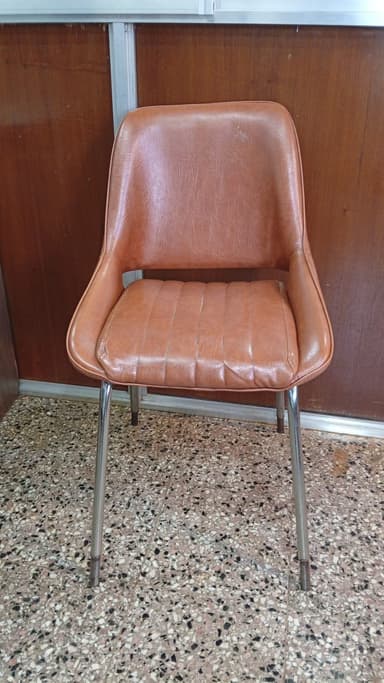 A modern dining side chair featuring a single-piece seat and back shell upholstered in tan faux leather, supported by four slender, tapered metal legs with a polished chrome finish. The seat cushion has horizontal channel tufting, and there is a small gap between the seat and backrest. — 3D model