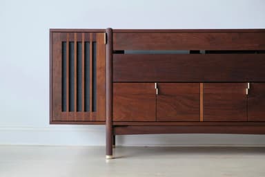 A long, low, dark wooden cabinet with a distinct mid-century modern design. It features slatted sections on the curved ends, a central open shelf, and multiple drawers and cabinet doors below, all adorned with small brass pull handles. The cabinet is supported by angled, tapered legs with brass ferrules at the bottom. — 3D model