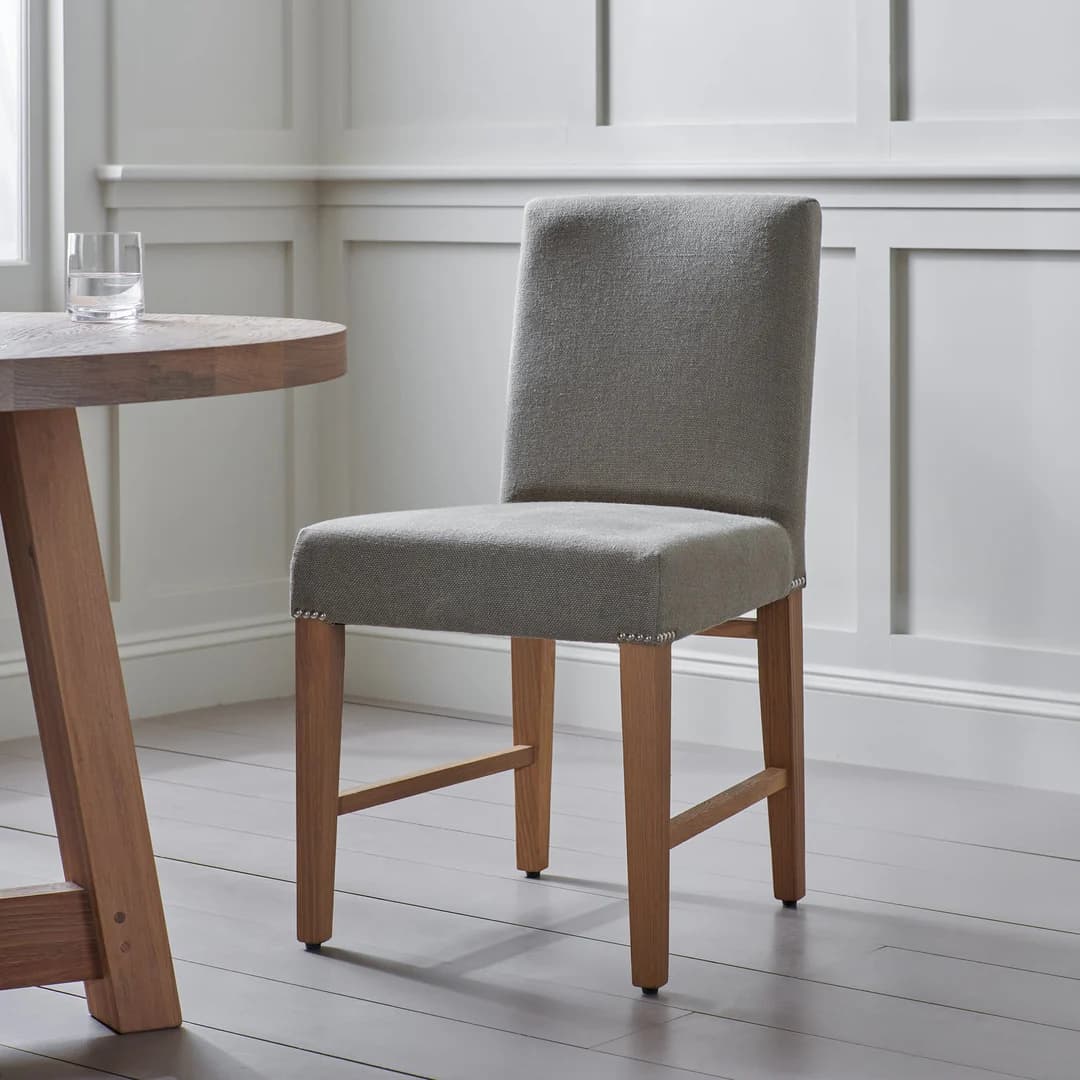 A high-backed upholstered chair with a square seat and four wooden legs. The upholstery is a textured fabric in a neutral grey tone. Decorative nailhead trim is visible along the edges of the seat and lower back support. The legs are made of wood with a light brown stain and feature a stretcher connecting the front and back legs on each side. — 3D model