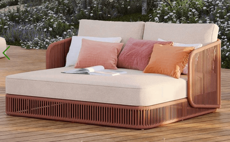 A contemporary daybed featuring a slatted light brown wooden frame with integrated armrests and back support. It is fitted with thick, beige upholstered seat and back cushions, accompanied by several throw pillows in varying pastel shades of pink, orange, and white. — 3D model