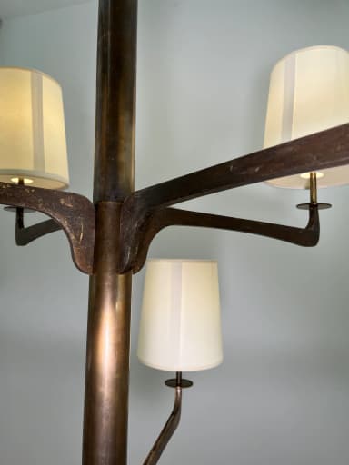 A modern multi-arm chandelier featuring a dark bronze-finished metal frame and eight tapered, drum-style fabric shades. The central column extends from a chain suspension, with eight arms branching out, each supporting an upward-facing light socket covered by a cream-colored textile shade. The metalwork exhibits a rustic or aged patina, suggesting a blend of modern design with traditional material treatment. — 3D model