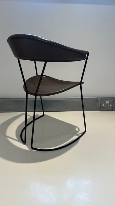 A contemporary armchair with a dark brown leather seat and backrest supported by a slender black metal frame. The frame features a curved base that extends into the legs, providing stability and a dynamic visual element. The seat and backrest are subtly contoured for comfort and appear to be constructed from stitched leather panels. — 3D model