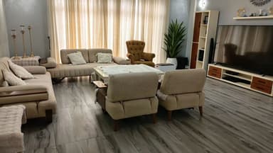 A modern sofa upholstered in a light beige fabric, featuring a distinctive channeled backrest and seat cushions. The arms are slightly curved with integrated wooden panels on top. The sofa appears to be composed of two separate seat sections and is supported by four dark, tapered wooden legs. It includes two decorative throw pillows matching the upholstery. — 3D model