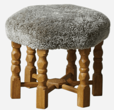 A small, round stool featuring a soft, shaggy grey upholstered seat and four turned wooden legs with stretcher supports. The legs have a light brown wood finish, and the overall design combines rustic and cozy elements. — 3D model