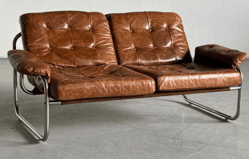 A two-seater sofa with a tufted seat and back, upholstered in brown leather. The frame is constructed from tubular metal, providing a contrasting industrial aesthetic. The armrests are also upholstered and integrated into the metal frame. — 3D model