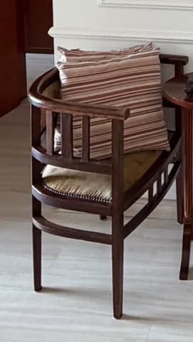 A compact armchair featuring a dark stained wooden frame with a distinctive barrel-shaped, curved backrest and open slatted sides. The chair has a firm, upholstered seat cushion in a light beige or cream color, accented by visible nailhead trim along the base. A separate, striped fabric pillow in shades of brown and beige rests against the backrest. The overall design suggests a blend of traditional and transitional aesthetics. — 3D model