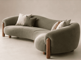 A long, curved sectional sofa with an organic, undulating shape, upholstered in a textured sage green fabric, supported by chunky, rounded wooden legs. — 3D model