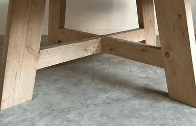 A round dining table with a thick, solid wood top and a substantial base constructed from heavy timber. The base features four angled legs connected by a cross-brace structure, providing stability and a rustic aesthetic. — 3D model
