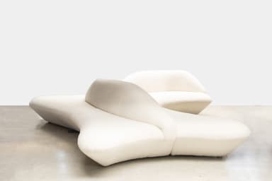 A modular sofa with an organic, curvilinear design, upholstered in a textured boucle fabric. The sofa appears to be composed of multiple connected units, creating an unusual, flowing silhouette that deviates from traditional sofa forms. The front section has a low profile with rounded edges, while the back section features a more upright, curved backrest. — 3D model