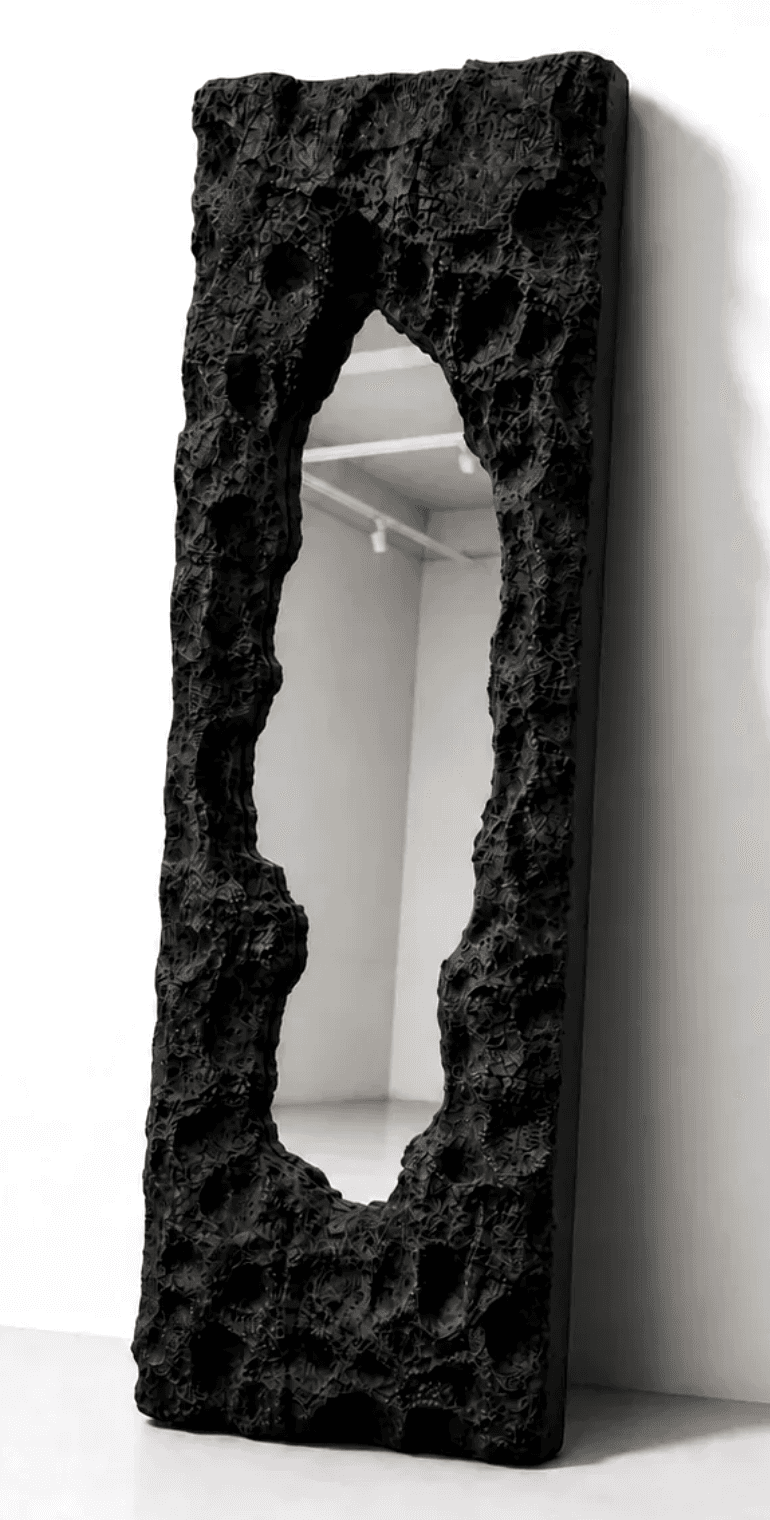 A tall, floor-standing mirror featuring a highly distinctive, irregular, and deeply textured black frame. The frame has an organic, almost volcanic or rock-like appearance, encasing a central mirror panel. The mirror itself is shaped with an abstract, non-uniform aperture, creating a visual void within the heavily sculpted surround. The object is leaning against a plain white wall, highlighting its artistic and dramatic form. — 3D model