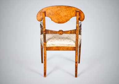 An antique armchair featuring a distinctive curved and carved backrest, likely made from curly maple or birch wood with a warm, amber finish. The chair has an upholstered seat covered in a cream-colored damask fabric with a patterned motif. The back legs are straight and square-tapered, and the back structure includes carved elements that function as armrest supports. The overall design suggests a formal, historical aesthetic. — 3D model