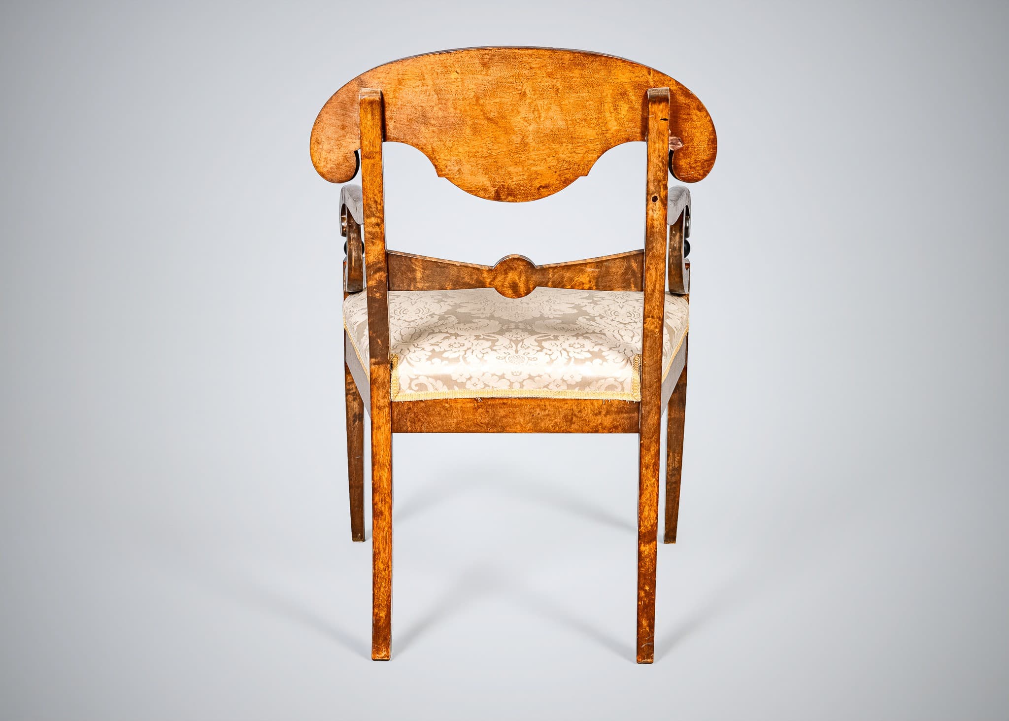 An antique armchair featuring a distinctive curved and carved backrest, likely made from curly maple or birch wood with a warm, amber finish. The chair has an upholstered seat covered in a cream-colored damask fabric with a patterned motif. The back legs are straight and square-tapered, and the back structure includes carved elements that function as armrest supports. The overall design suggests a formal, historical aesthetic. — 3D model