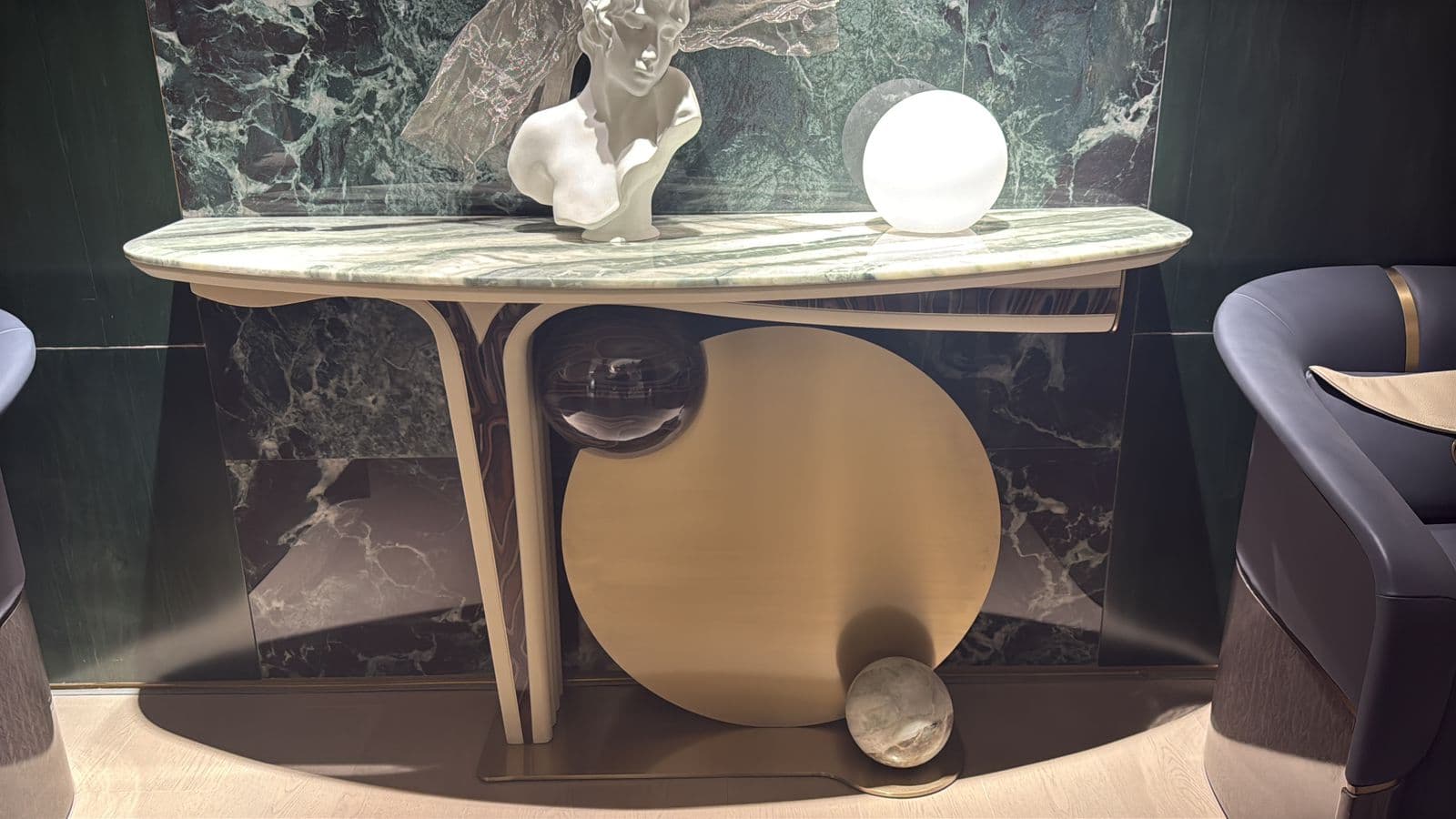 An elegant and sculptural console table featuring a grey and green veined marble top with an asymmetrical, curved edge. The base is crafted from polished gold-toned brass, forming a dynamic composition of geometric shapes, including a large circular disc, vertical ribbed elements, and a small marble sphere at its base, creating a distinctive Art Deco-inspired aesthetic. — 3D model