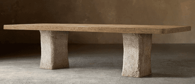 A rectangular coffee table featuring a thick, light-toned wood plank top with visible grain, supported by two substantial, roughly textured stone or concrete block legs. The design emphasizes natural materials and a minimalist, robust aesthetic. — 3D model