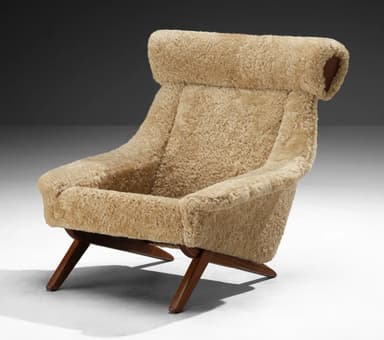 A high-backed armchair with a distinctive sloped silhouette. The chair is upholstered entirely in a textured, light brown sheepskin or shearling material, giving it a plush and organic appearance. The seat and back cushions are integrated into the overall form. The base consists of dark wood legs with a visible grain, angled outwards for stability. The headrest area is pronounced and rolls over the top of the backrest. — 3D model
