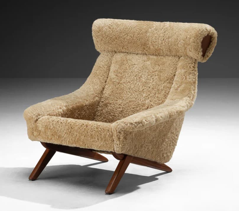 A high-backed armchair with a distinctive sloped silhouette. The chair is upholstered entirely in a textured, light brown sheepskin or shearling material, giving it a plush and organic appearance. The seat and back cushions are integrated into the overall form. The base consists of dark wood legs with a visible grain, angled outwards for stability. The headrest area is pronounced and rolls over the top of the backrest. — 3D model