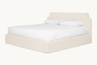 A bed frame with a padded headboard and platform base, covered in a neutral-toned fabric. The headboard has a distinctive silhouette with curved upper corners. The bed is shown with white linens and two pillows. — 3D model