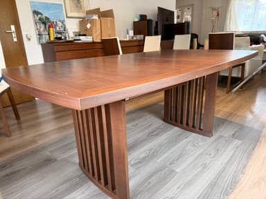 A modern dining table featuring a dark brown wooden top with elegantly curved edges and a matching slatted base. The table top displays a visible wood grain pattern, suggesting a veneer finish, and shows some minor surface wear. The base appears robust, designed with vertical wooden slats, which likely form a pedestal or trestle-style support. — 3D model