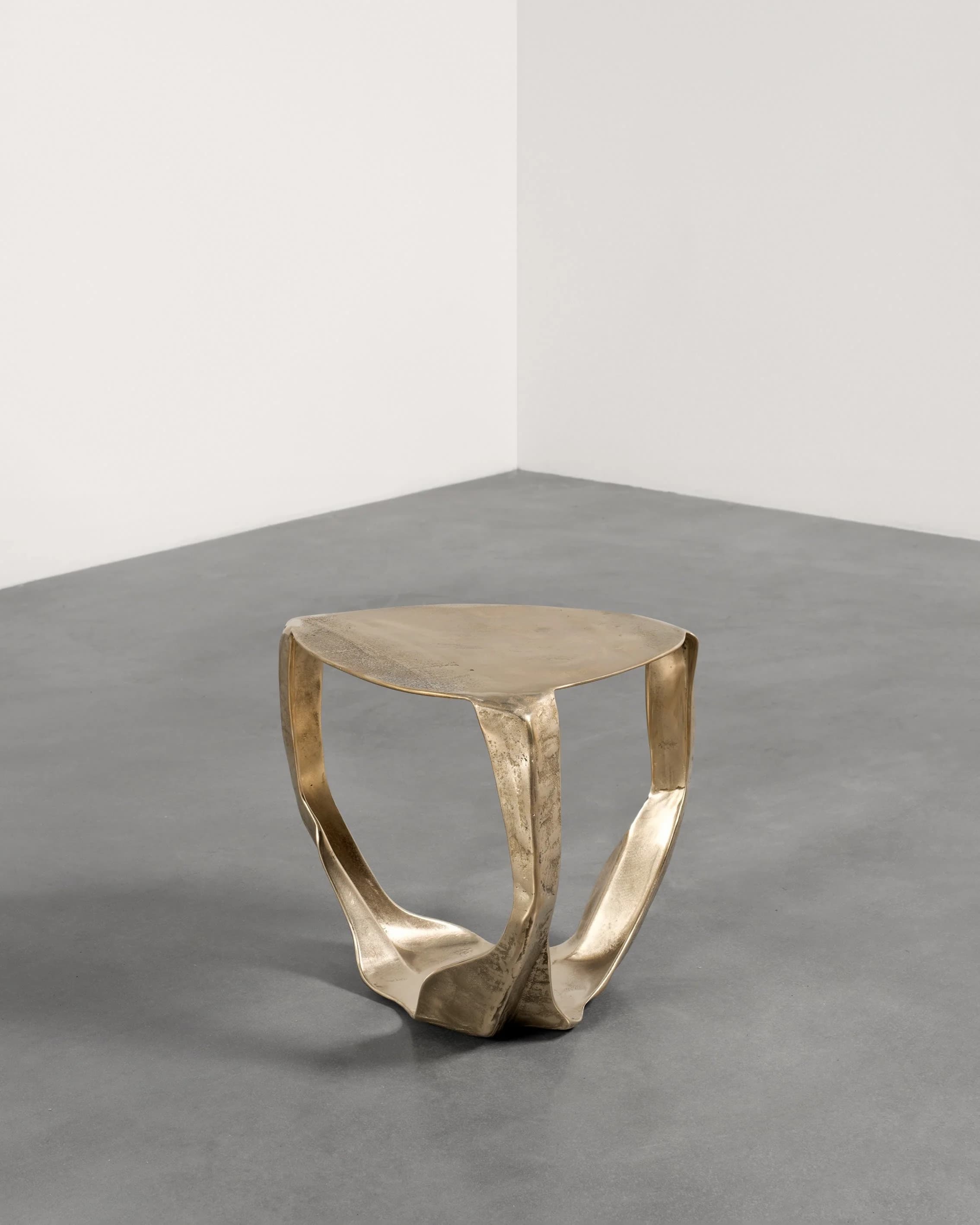 A sculptural accent table crafted from brass with an organic, fluid form. The table features a rounded triangular top and a base composed of three sinuous, interconnected legs that converge towards the ground, creating a dynamic and artistic silhouette. The brass exhibits an antiqued or distressed finish, enhancing its visual texture and depth. — 3D model