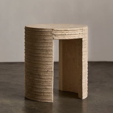 A cylindrical end table with a textured, stone-like surface. The table has a rounded top and a single, curved leg or base element visible. The side of the table exhibits a series of horizontal grooves or ridges, suggesting a layered construction or decorative carving. The material appears porous and natural, consistent with travertine or a similar stone composite. The color is a light, earthy beige. — 3D model